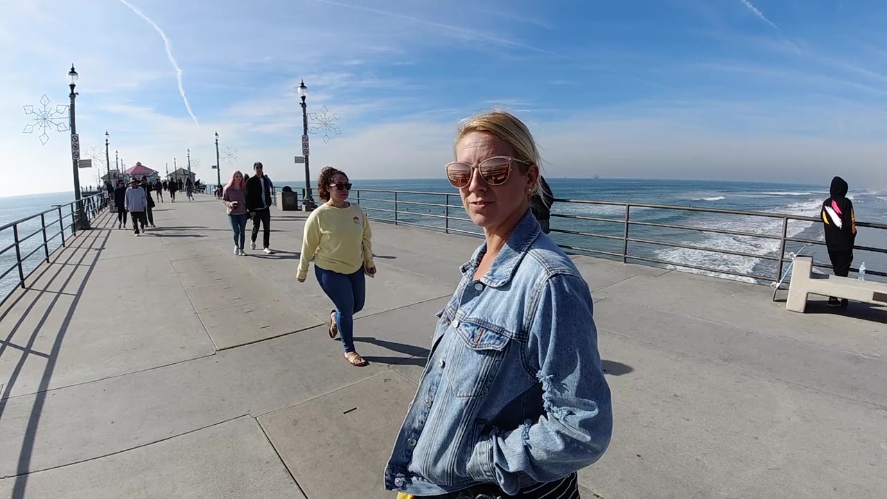 At 0:02, the woman in the denim jacket is framed closer at right on the Huntington Beach pier, with the ocean horizon, white surf, and several walkers in the background.