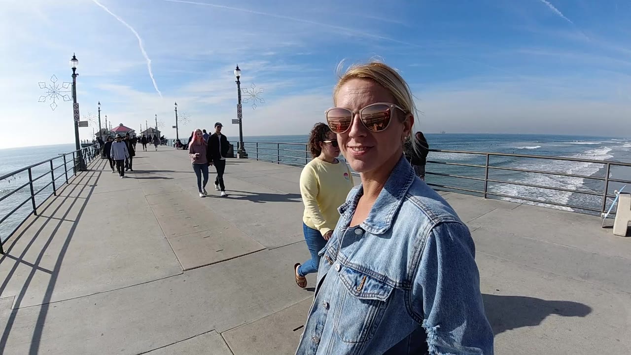 Woman in sunglasses near the camera on a pier while people walk toward the ocean view.