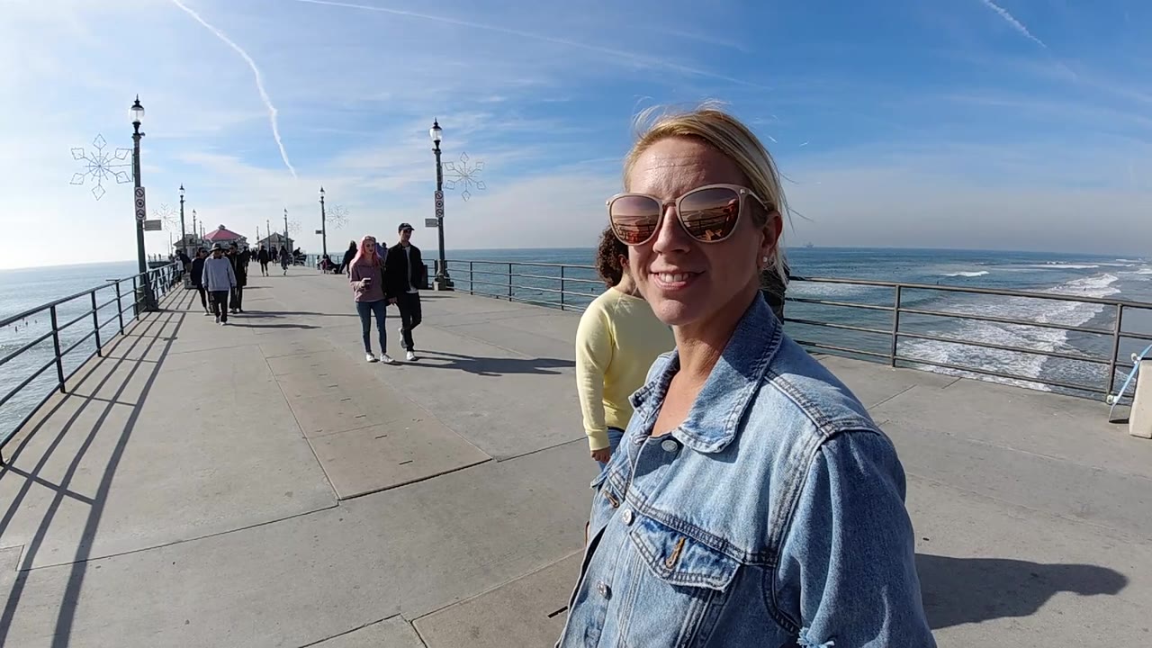A close right-side view shows the smiling woman on the Huntington Beach pier, with the ocean and breaking waves to the right and small groups of pedestrians continuing ahead.