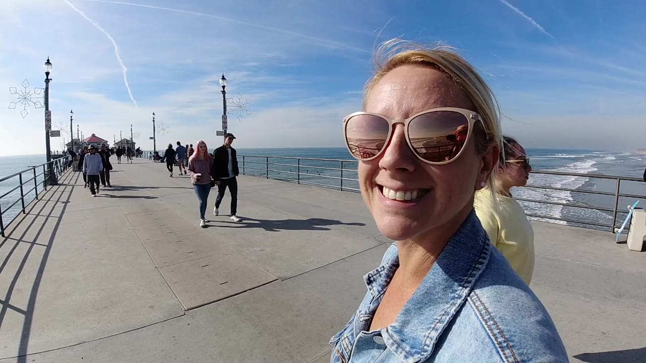 At 0:01, the woman remains in the right foreground on the Huntington Beach pier as walkers approach along the concrete path and the surf line runs behind the railing.