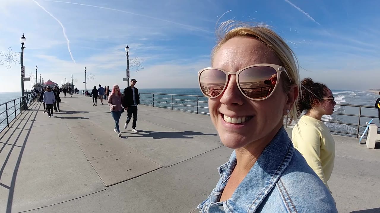 At 0:02, the smiling woman fills the right side of the frame, while a small group of people walks down the Huntington Beach pier beside the ocean under bright blue sky.