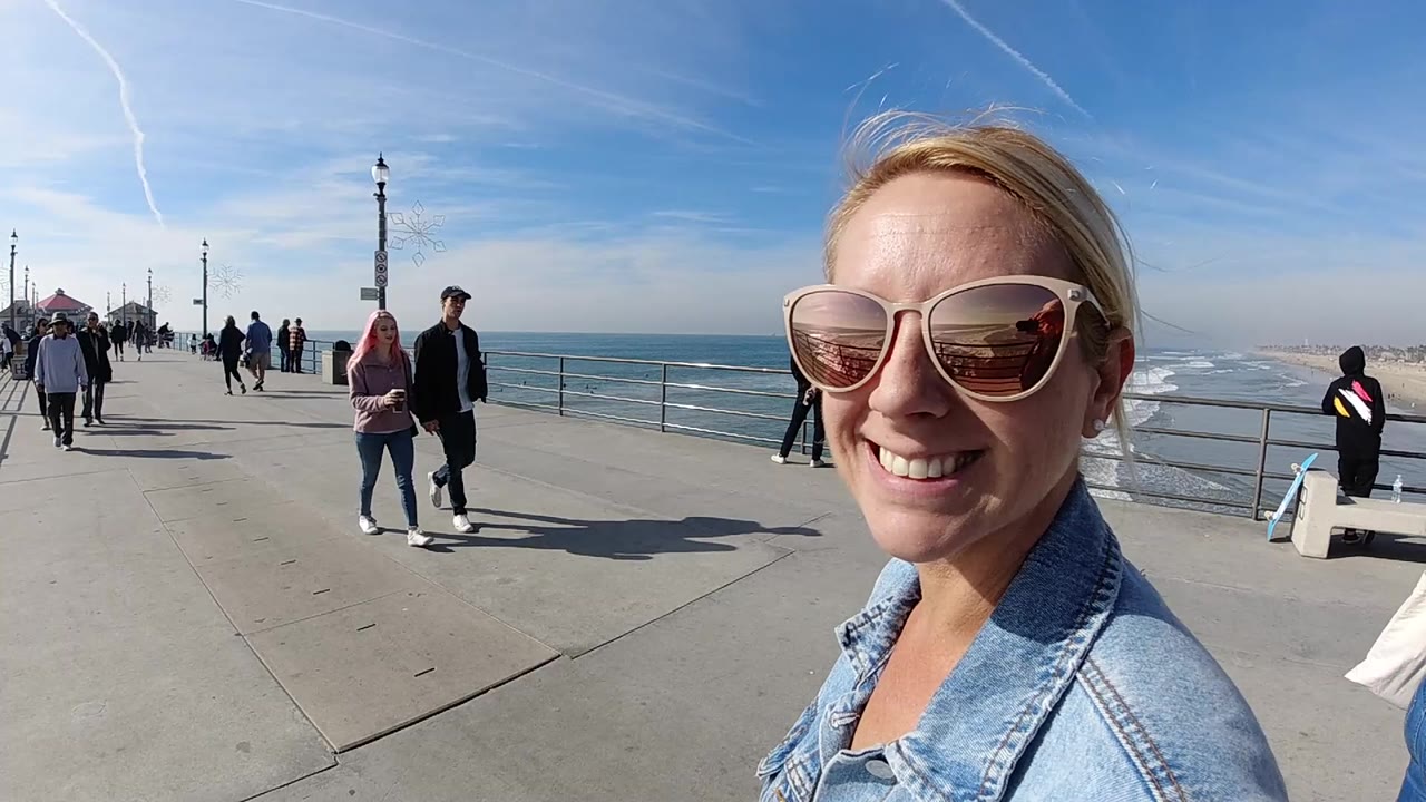The woman remains close in the right foreground at Huntington Beach, smiling in sunglasses while walkers, a railing, the ocean horizon, and bright sky fill the background.