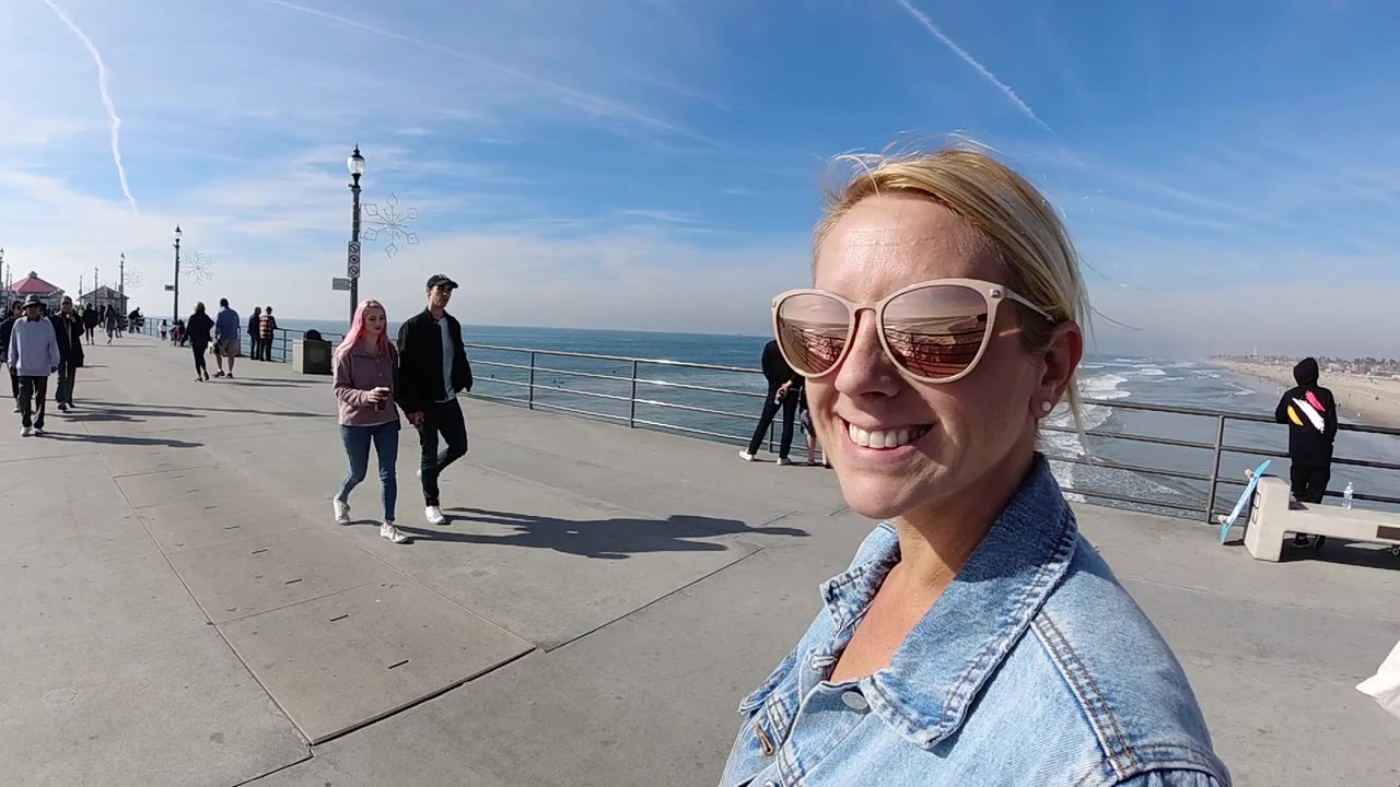 At Huntington Beach, the smiling woman shifts toward the right edge while waves, sandy shoreline, a bench, railing, and several pedestrians remain visible under clear daylight.