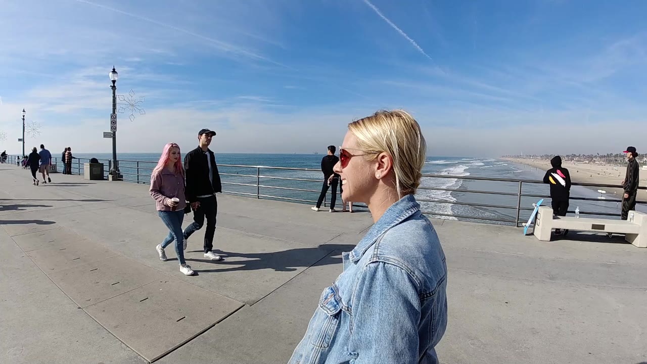 Woman in denim jacket near pier railing as pedestrians walk past the ocean.
