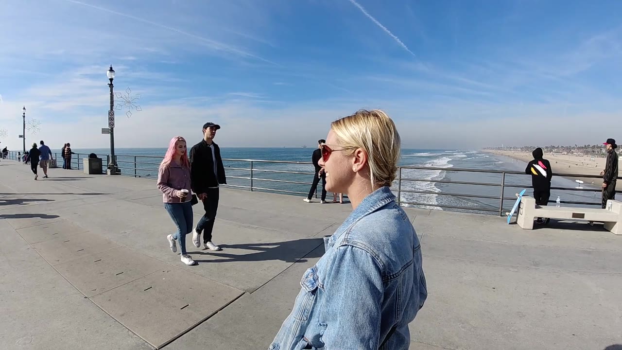 At 0:02 in Huntington Beach, the woman in denim remains near the right edge as two pedestrians pass closer to the camera, with a seated figure by a bench and the beach stretching into haze behind the railing.