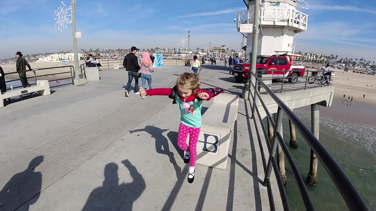 A young girl jumps near a concrete HB bench on the Huntington Beach pier, with railings, water below, pedestrians, and a red vehicle in the background.