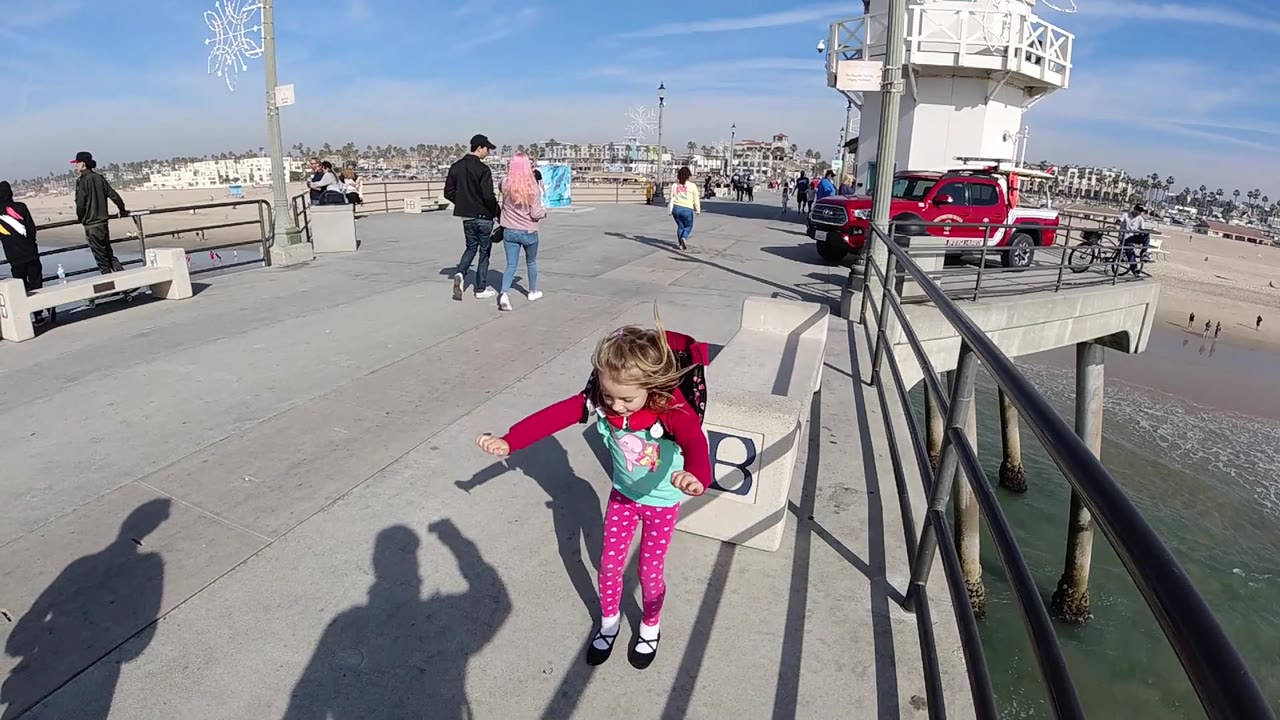 The girl looks down beside the HB bench on the Huntington Beach pier, with the water edge, railings, parked red vehicle, bicycles, and distant beach buildings visible.