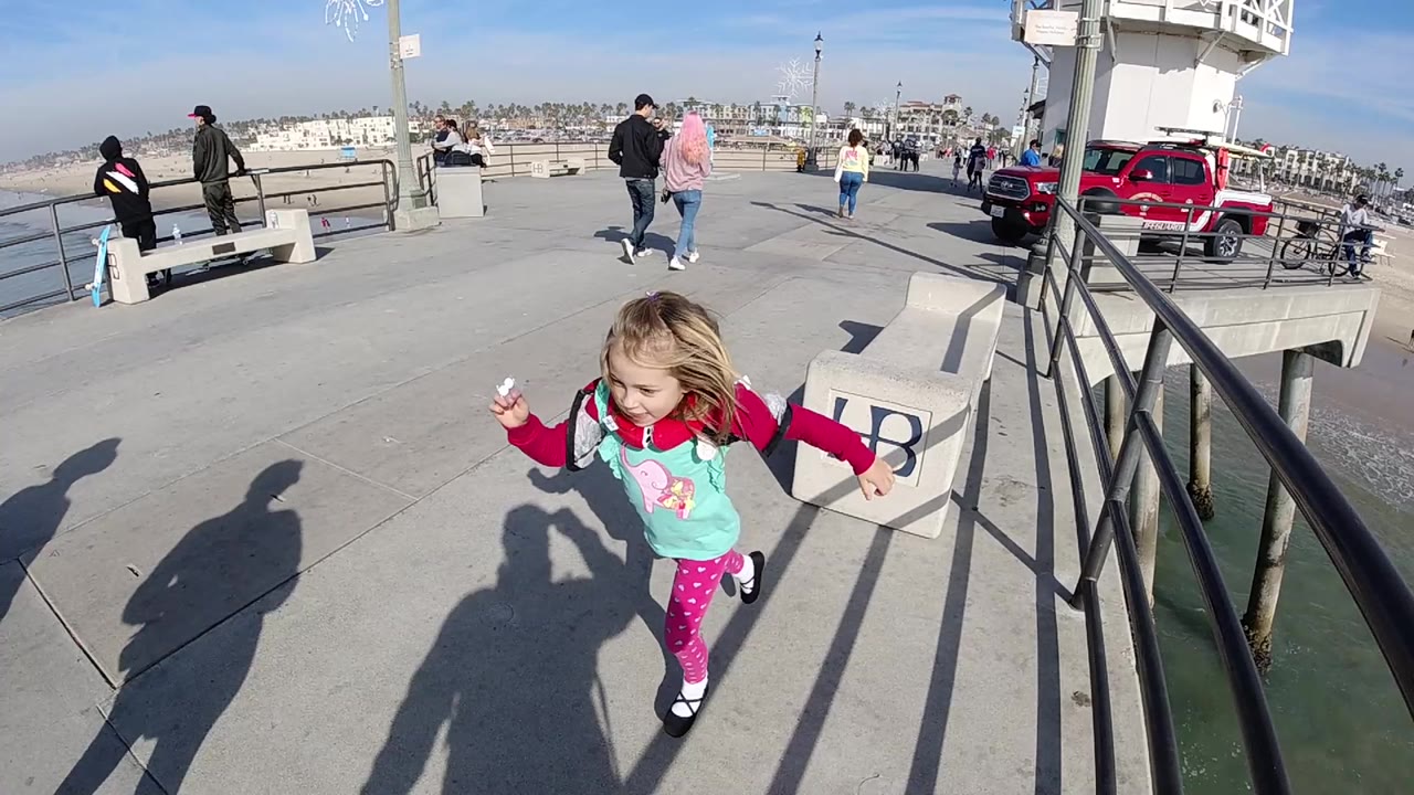 Girl in bright clothing moving across a pier walkway with ocean and benches nearby.