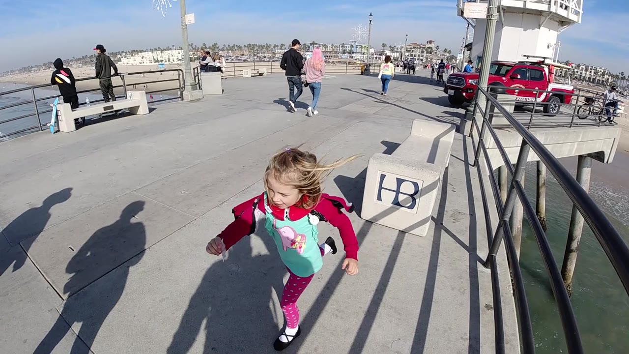 A young girl turns to the side in the foreground of the Huntington Beach pier, with the ocean edge, black railings, benches, pedestrians, and parked vehicles behind her.