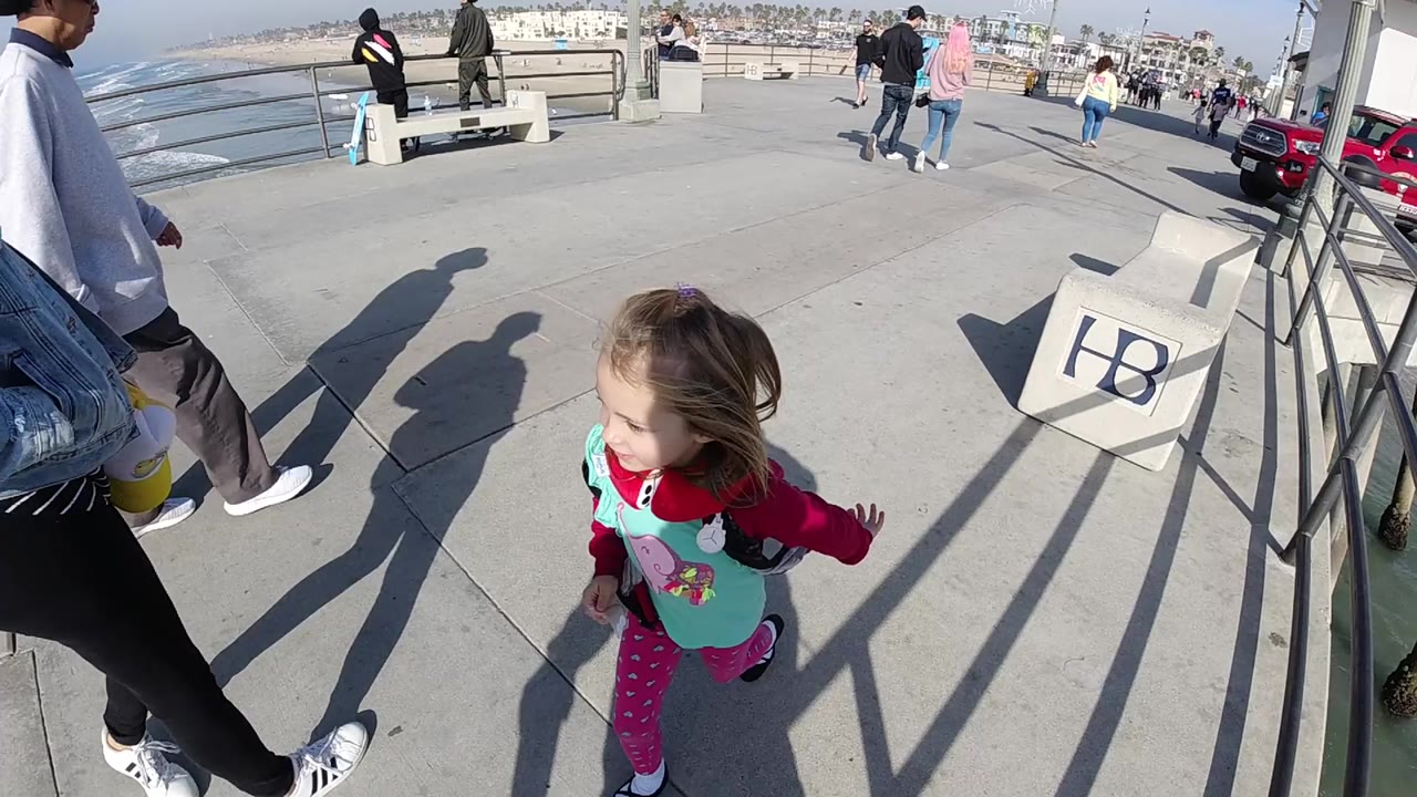 A young blonde girl in pink leggings runs along a Huntington Beach boardwalk as adults pass beside metal railings with ocean water visible to the left.