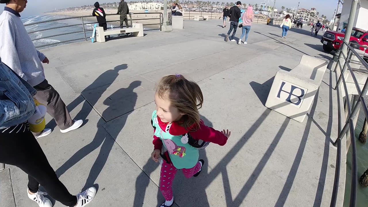 The girl continues running near the boardwalk railing in Huntington Beach, with pedestrians ahead, ocean water at the edge, and strong sun casting long shadows on the concrete.