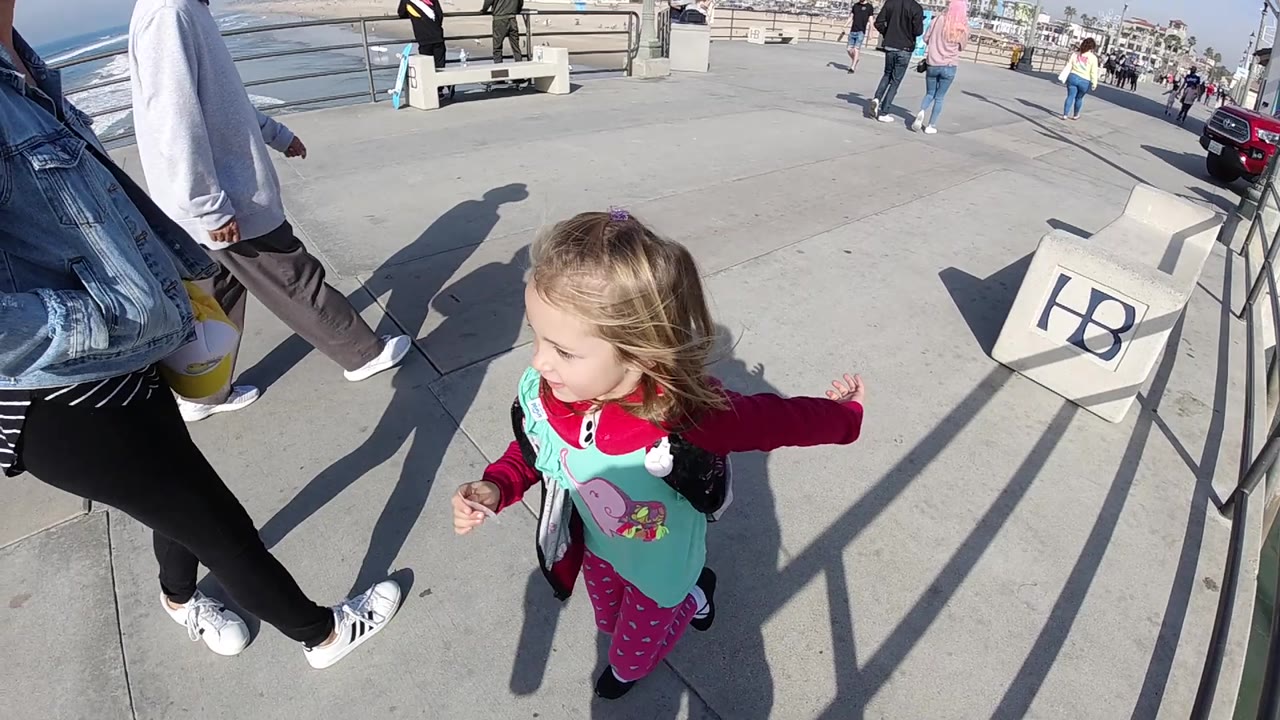 The child runs closer to the railings and a concrete sign marked with letters, with adults cropped at the edges and beach pedestrians visible in the distance in Huntington Beach.