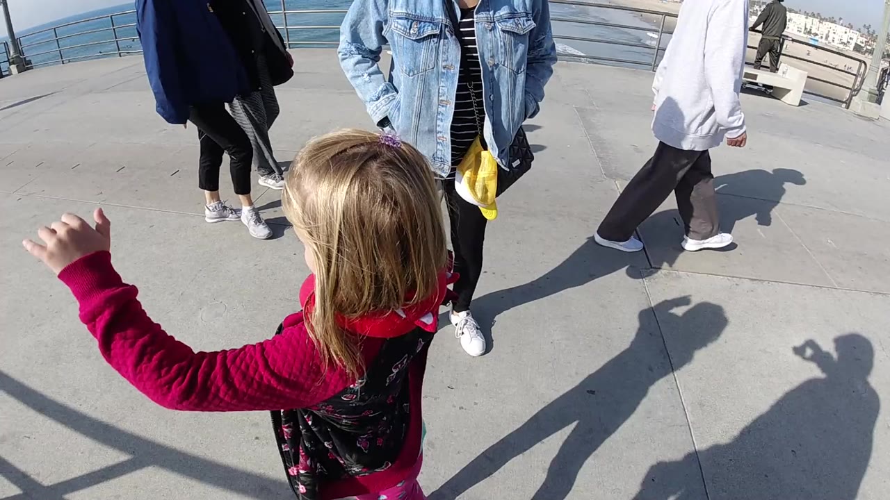 A little girl in a pink jacket is seen from behind on a Huntington Beach waterfront walkway, with adults nearby, metal railings, ocean water, and long shadows in bright sun.