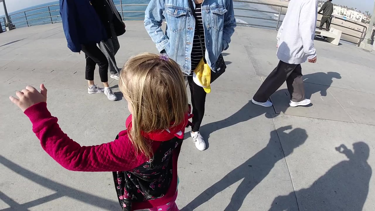 Girl raises one arm while adults stand and walk near the ocean railing.