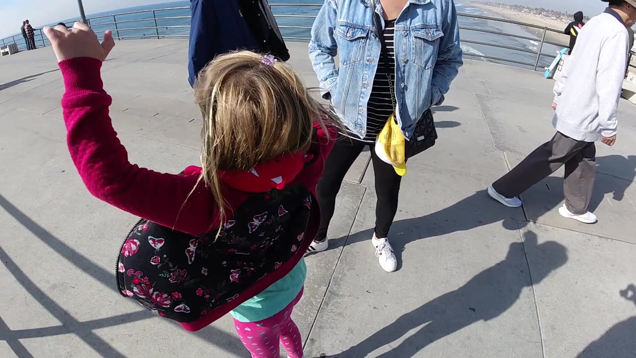 A rear view catches the little girl mid-jump in Huntington Beach, her pink jacket lifted by motion, with adults standing nearby and the ocean visible beyond the railing.