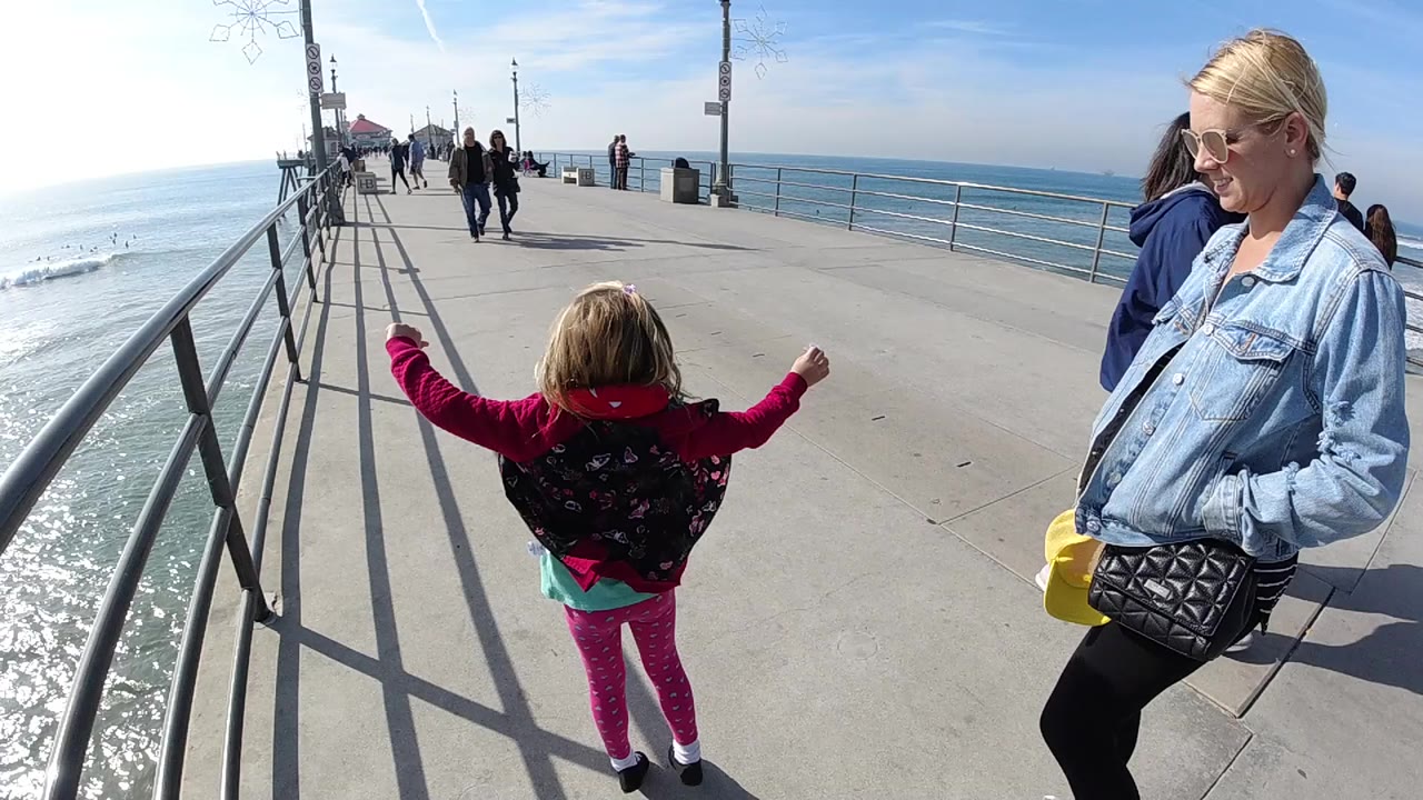The girl remains near the center of the Huntington Beach pier with both arms out, with the woman at right, railings leading forward, and sparkling water at left.