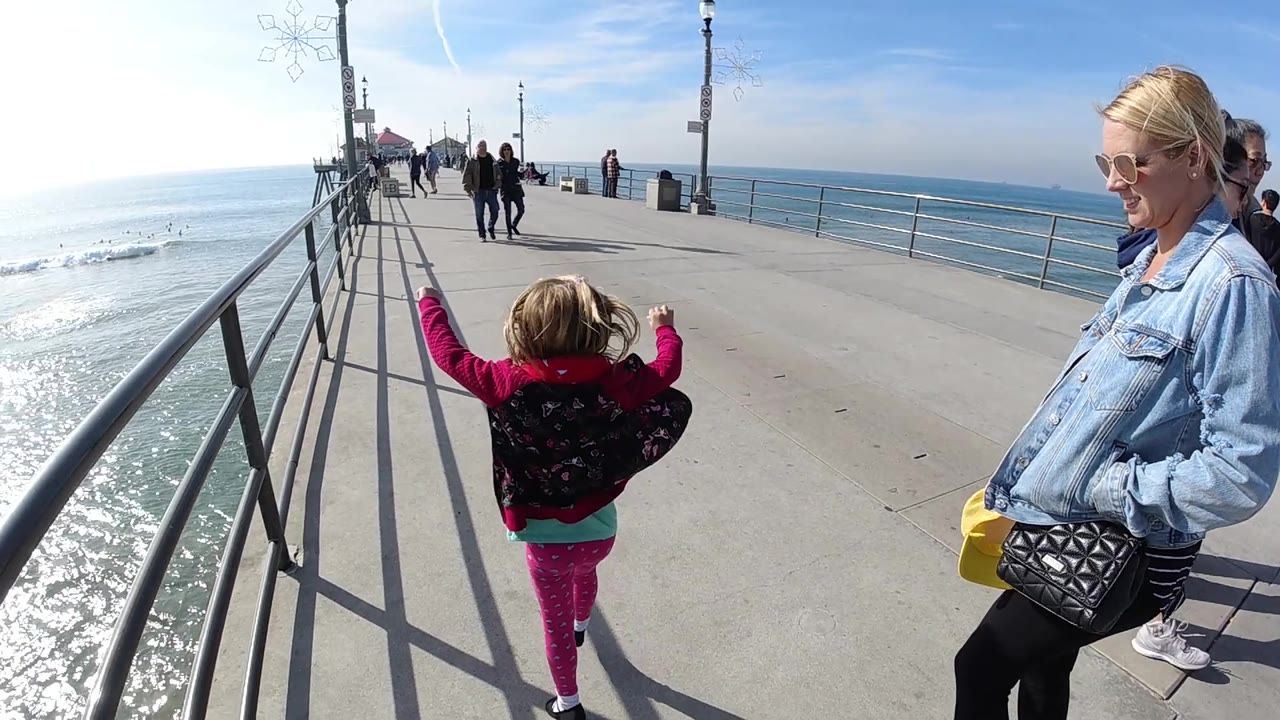The girl begins moving forward on the Huntington Beach pier with one arm lifted, while the woman stands at the right edge and the ocean horizon fills the background.