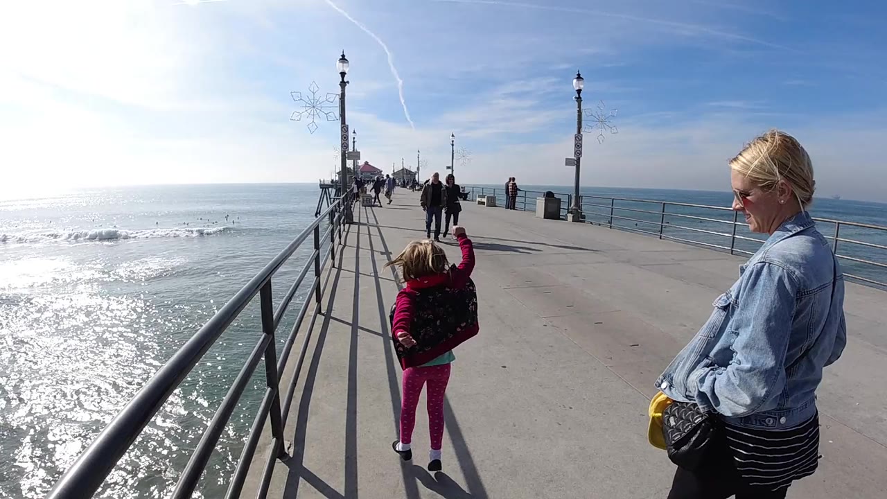 In Huntington Beach, a blonde woman in a denim jacket and a child in pink walk along a concrete pier with railings, lamp posts, ocean glare, and distant pedestrians ahead.