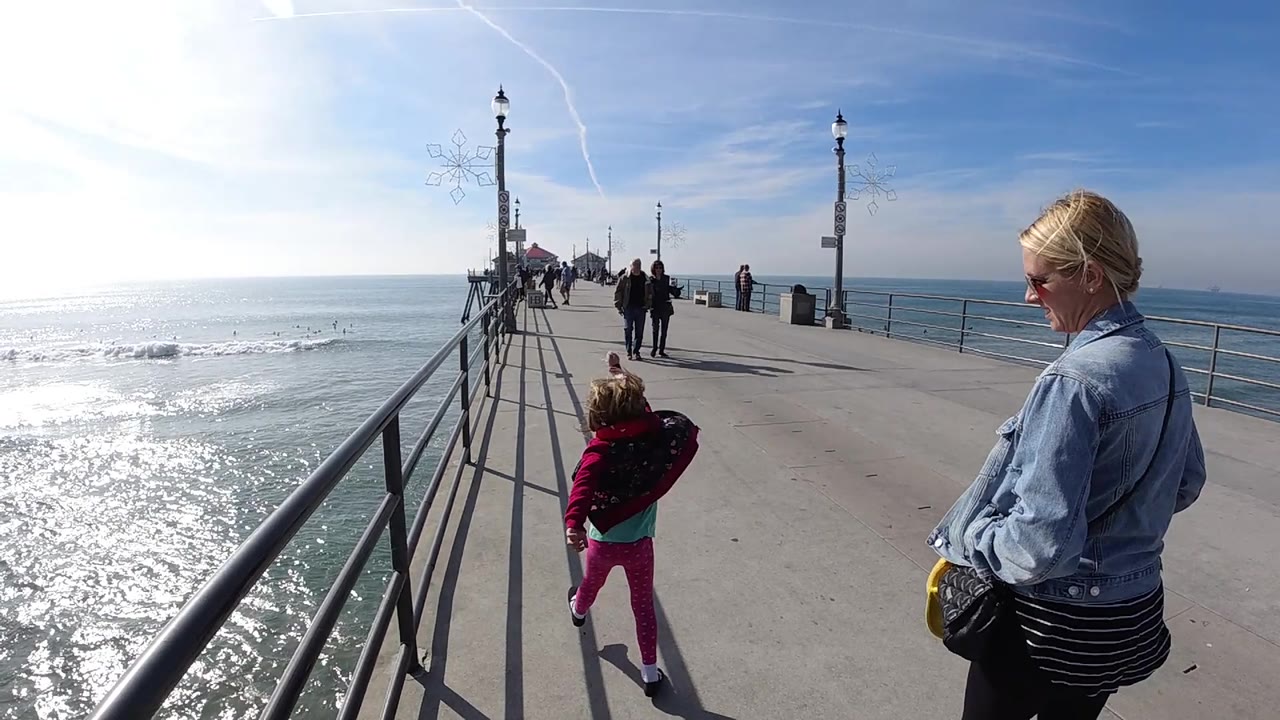 The woman stays near the right edge while the child moves farther down the pier, with Huntington Beach ocean water, railings, lamp posts, and pedestrians visible under clear blue sky.