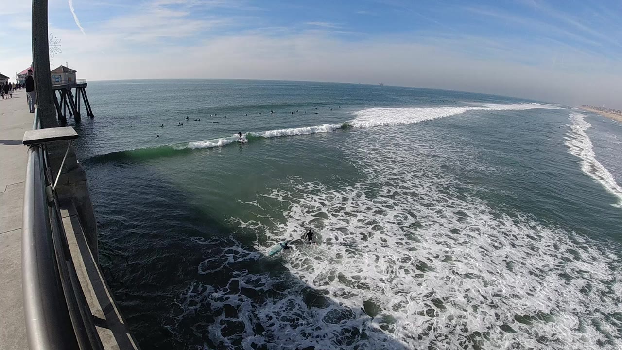 High view from a Huntington Beach pier railing over white surf, small surfers in the water, sandy shoreline at right, and no close visible faces.