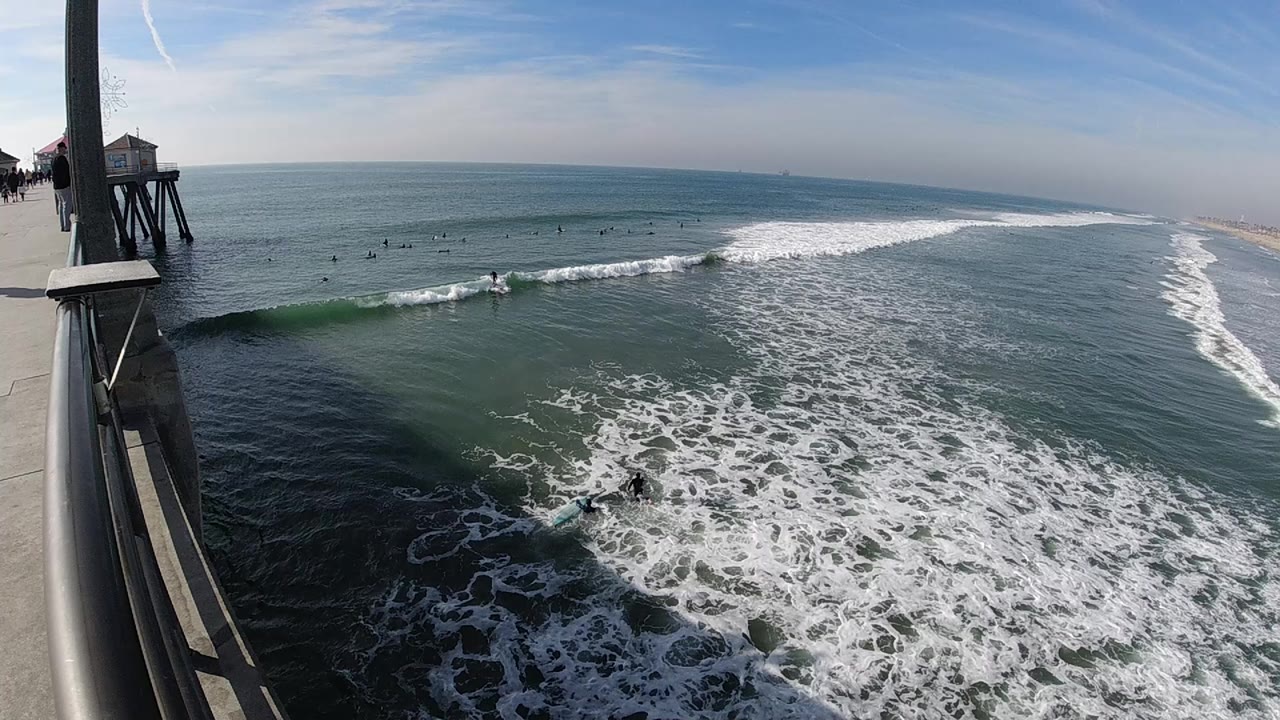 Huntington Beach ocean view with the pier edge on the left, a clean breaking wave across the center, surfers offshore, and pale beach stretching into the distance.