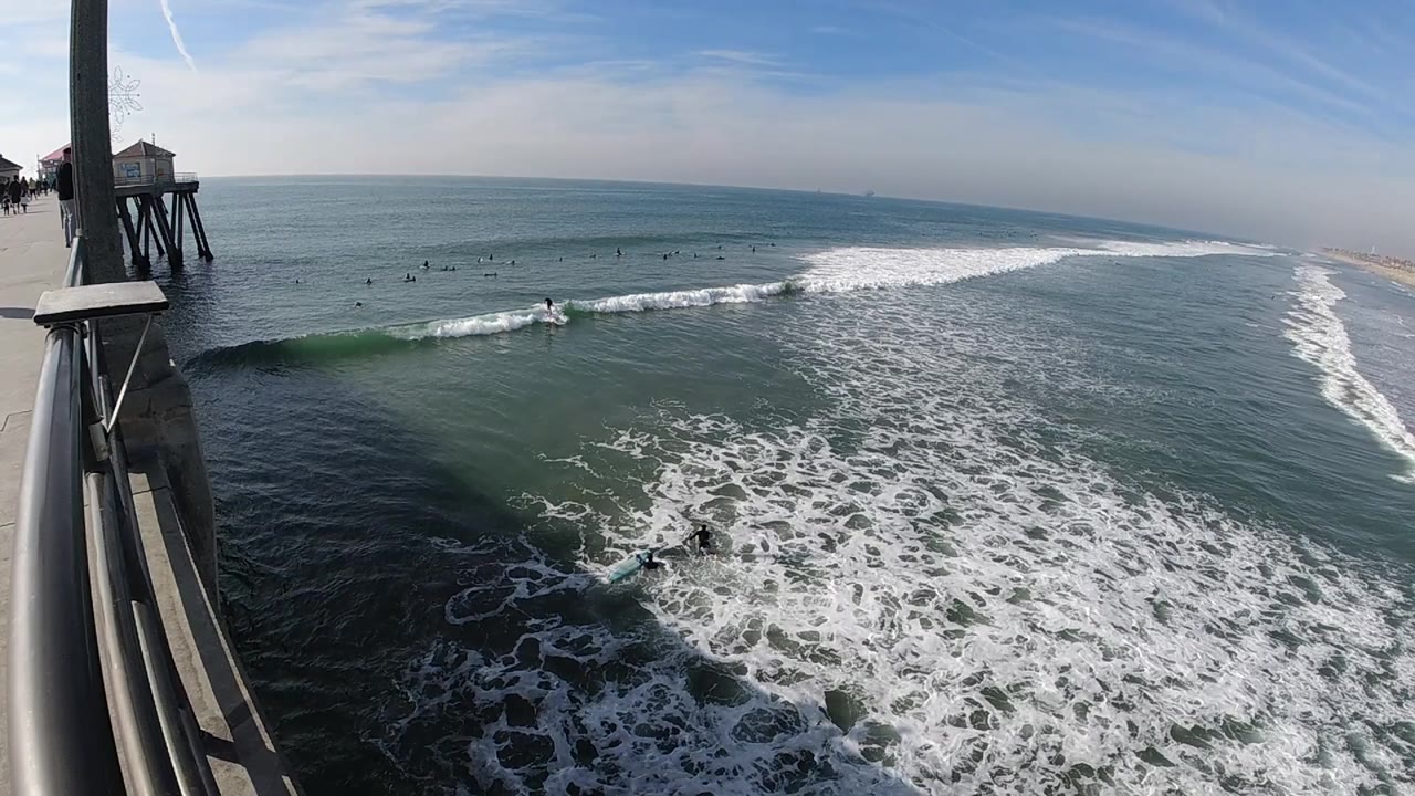 From beside the Huntington Beach pier structure, foamy surf spreads across the lower frame while small surfers wait beyond the breaking wave line.