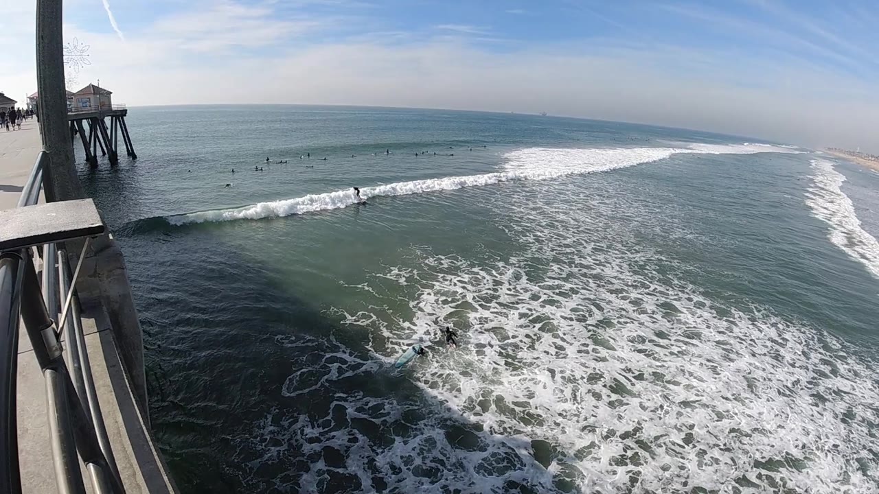 The Huntington Beach pier edge frames the left side while whitewater fills the lower frame and scattered distant surfers sit beyond the wave.