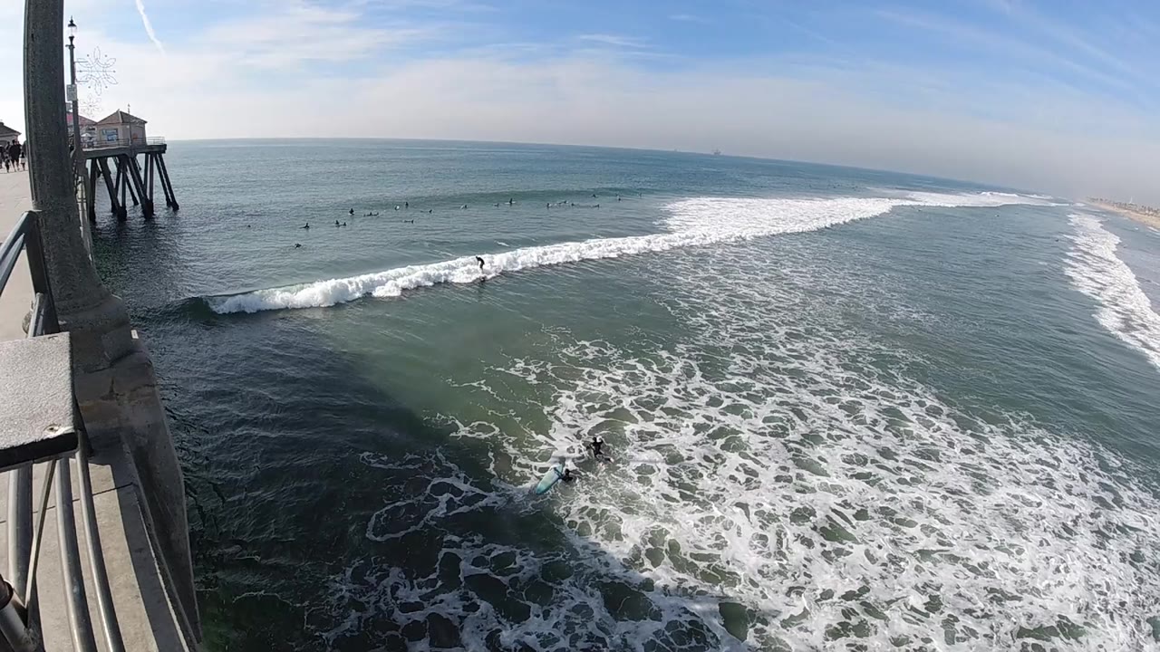 Huntington Beach surf fills the frame as a white wave band moves across the midground beside the left-side pier, with no visible people.