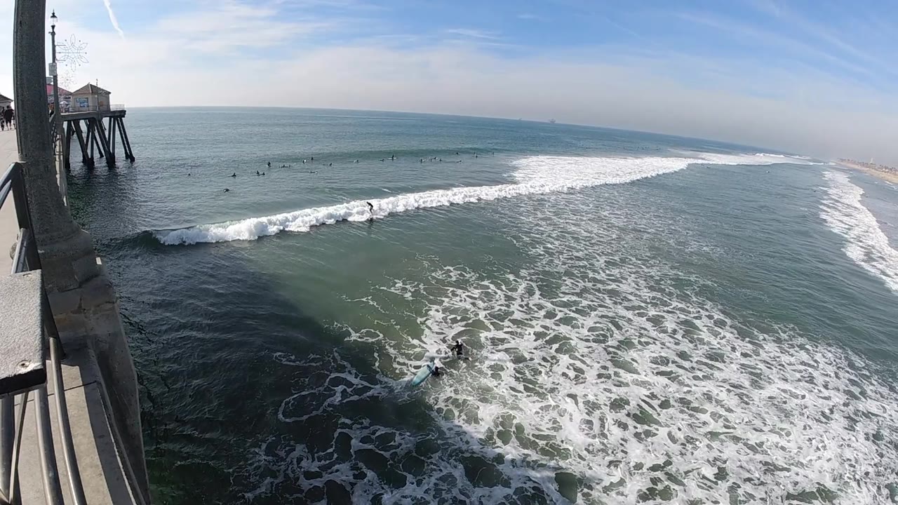 The pier remains at the left edge in Huntington Beach while whitewater spreads farther across the lower right water, with no visible people.
