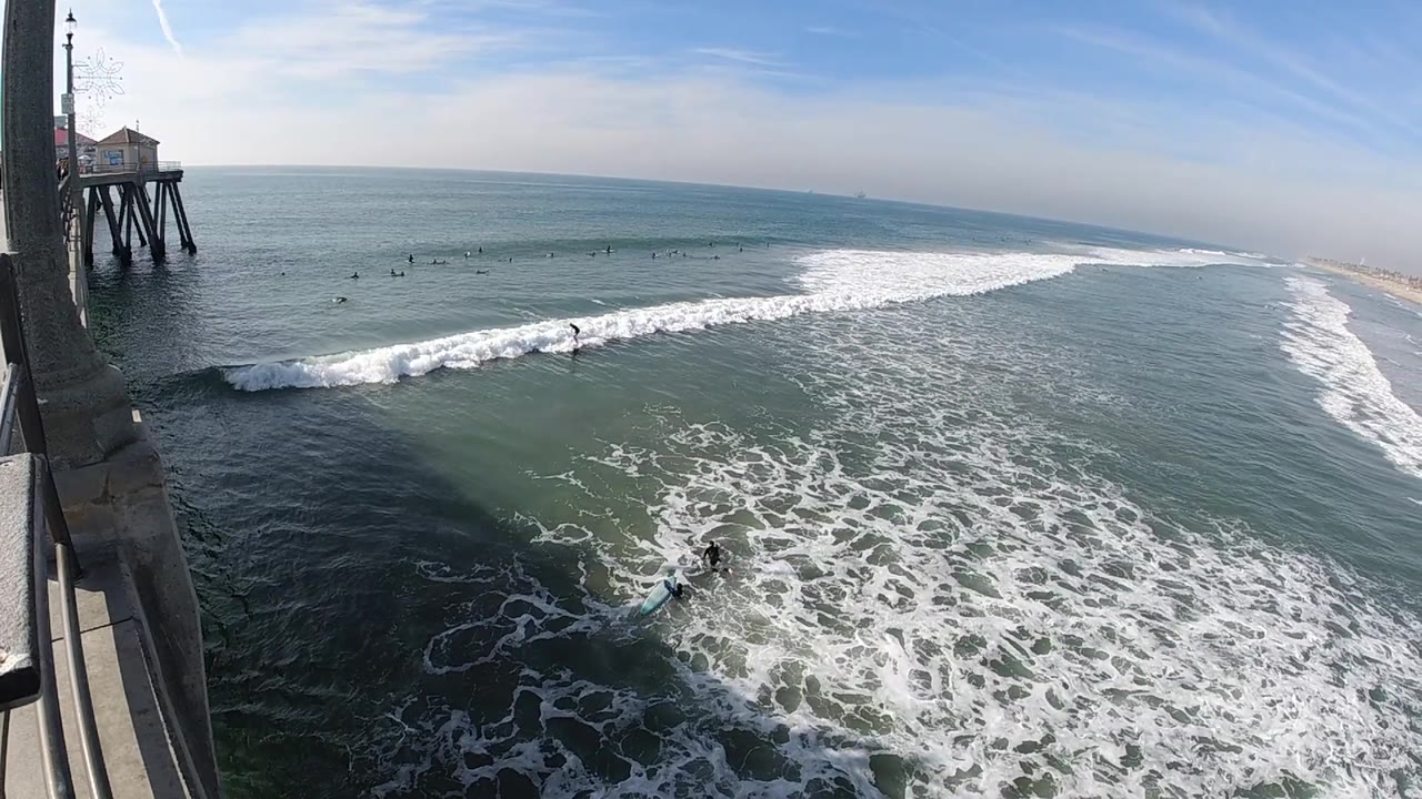 A breaking wave curls through the midground below the left-side pier at Huntington Beach, with foamy water in front and no visible people.