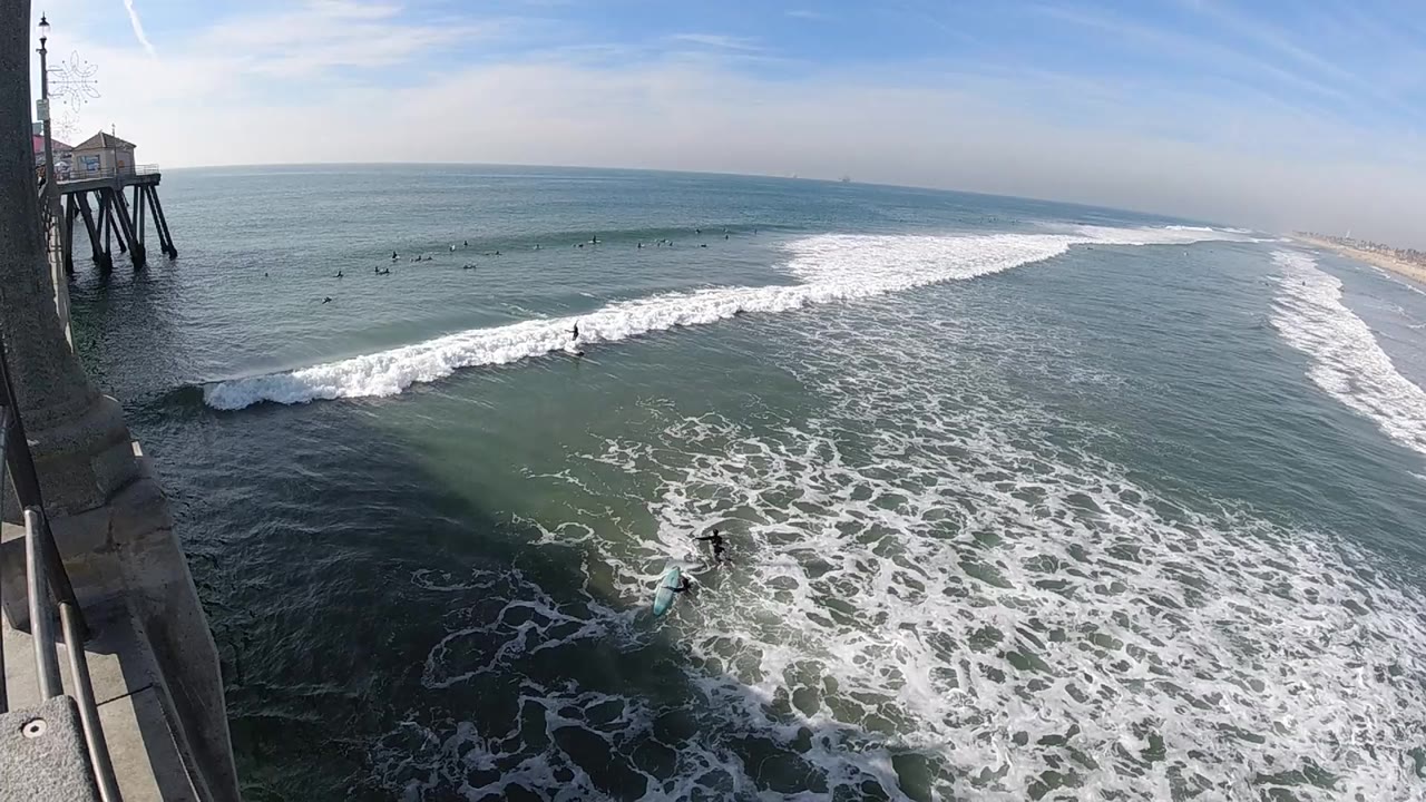 Elevated Huntington Beach view with a white wave breaking across blue-green water, pier structure at the left edge, distant shoreline to the right, and no visible people.
