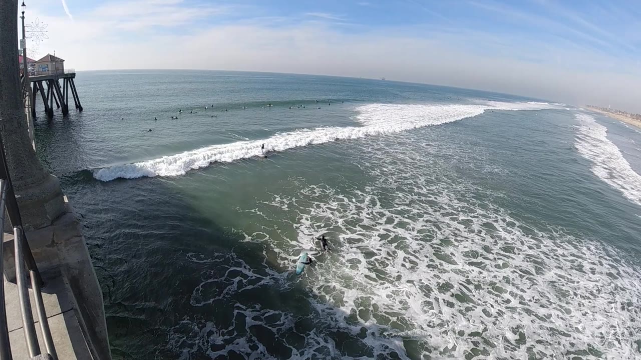 Huntington Beach surf scene with a long white breaker stretching across the center of frame, pier supports on the left, open ocean beyond, and no visible people.