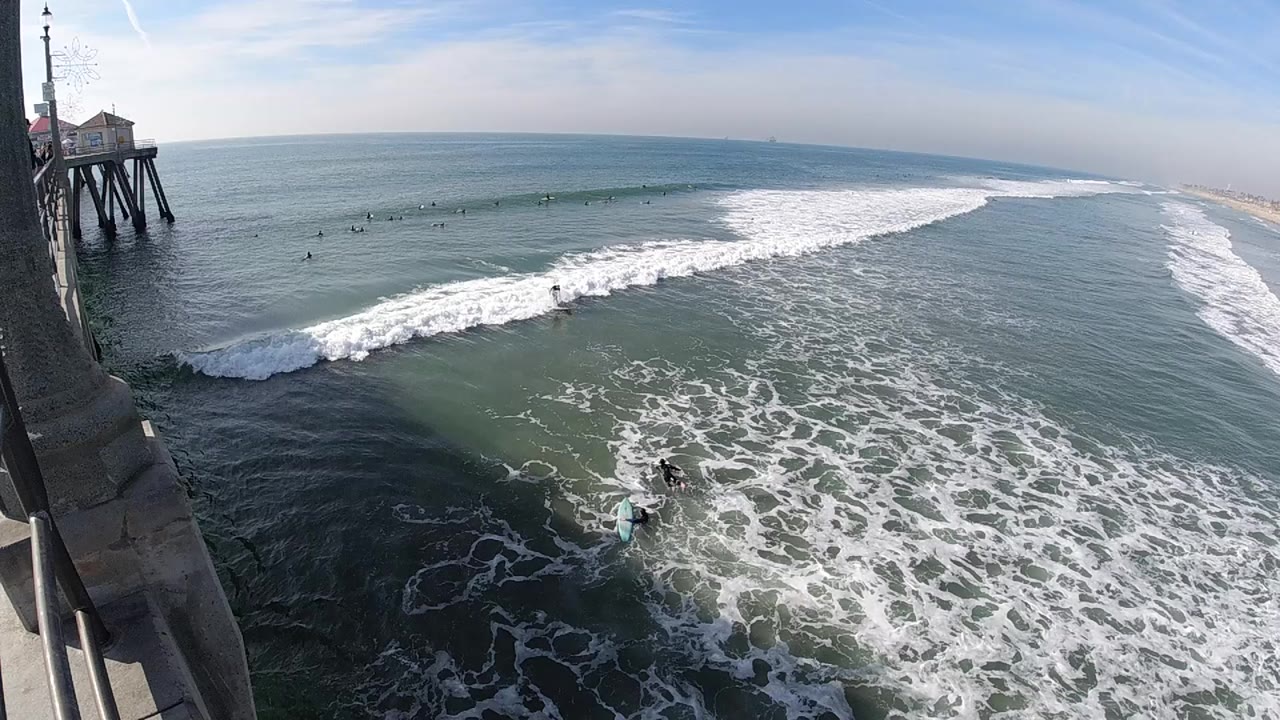 Huntington Beach surf rolls diagonally across the ocean while small figures wait in the water below the left-side pier structure.