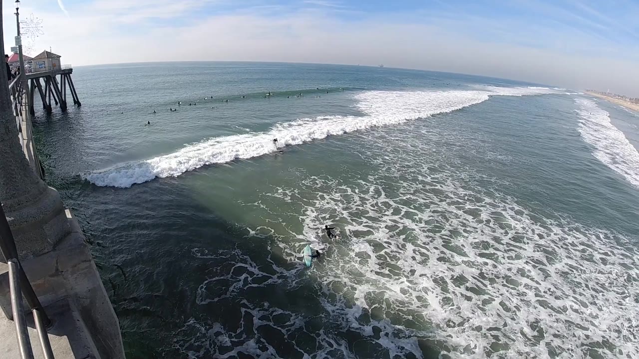 Huntington Beach water fills the frame as a white breaking wave advances toward the lower foreground and distant surfers sit beyond it.