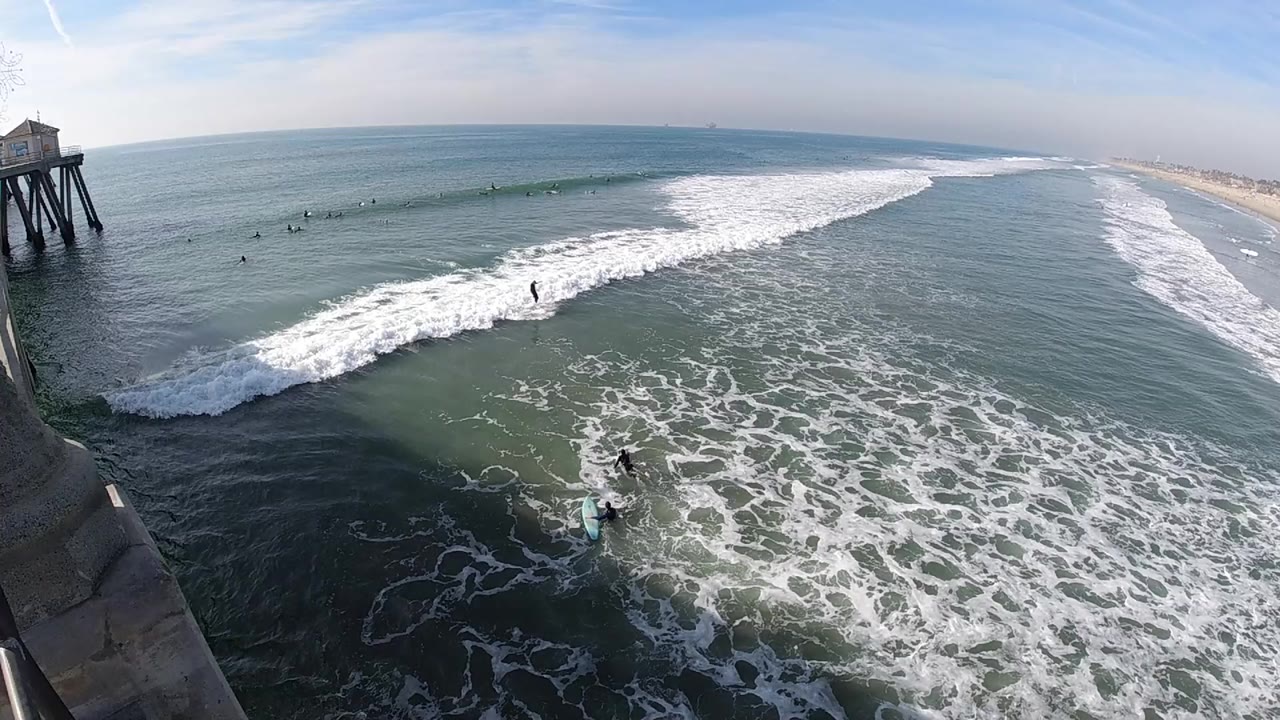 Elevated Huntington Beach view with pier supports at the left edge, white surf breaking across blue-green water, surfers visible offshore, and the sandy coastline fading into the distance.