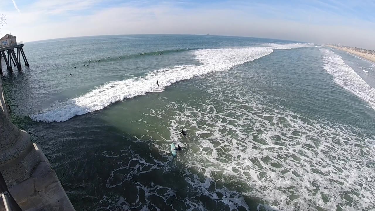 A broad Huntington Beach water view shows a larger sheet of foam in the foreground, surfers spaced across the midground, the pier on the left, and the long beach receding at right.