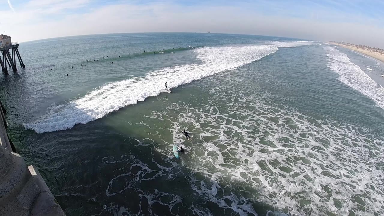 Huntington Beach surf rolls diagonally through the frame, with a larger whitewater band across the middle, scattered surfers beyond it, and the pier supports near the left edge.