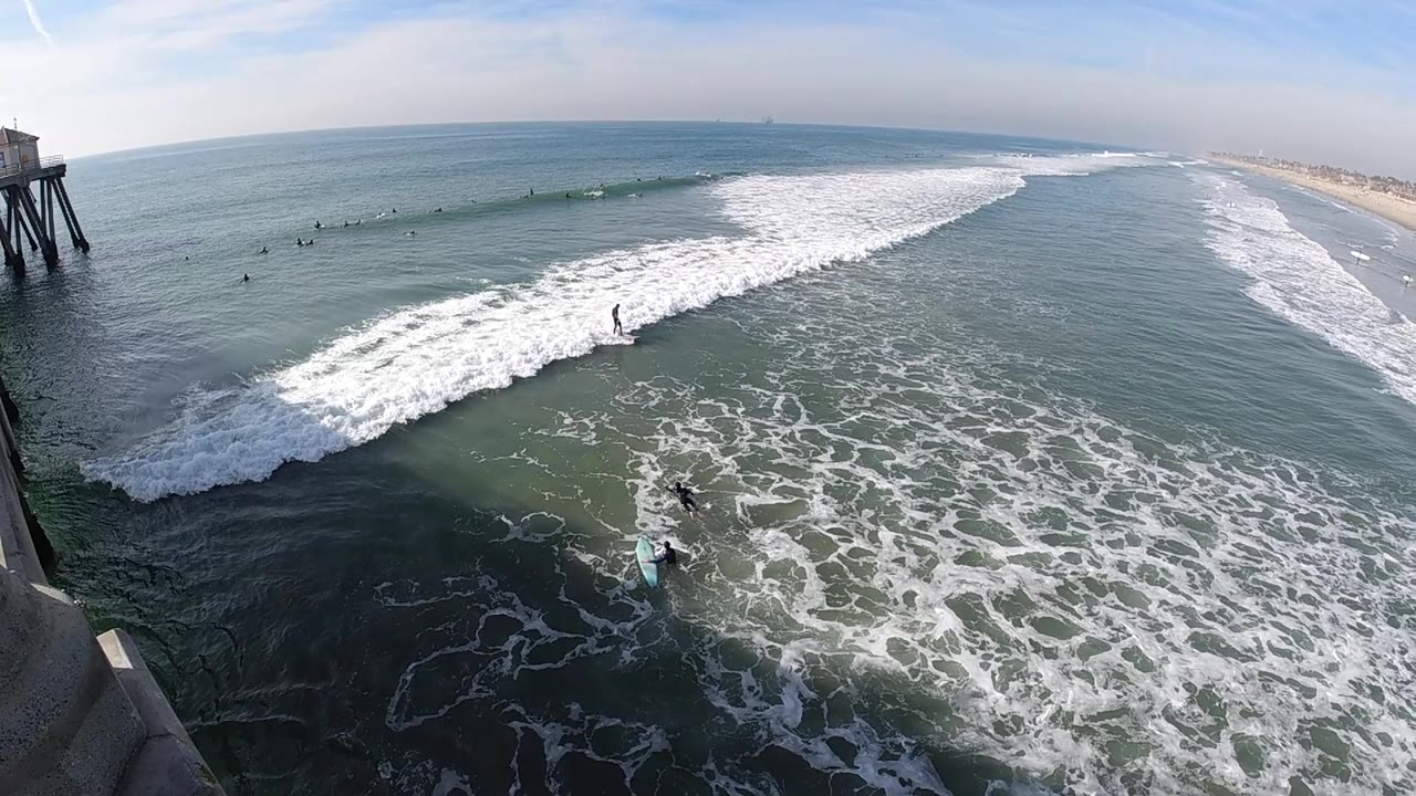 The pier remains at the left while a broad white wave spreads toward the lower frame, with surfers visible as small dark figures in the calmer water beyond the break.