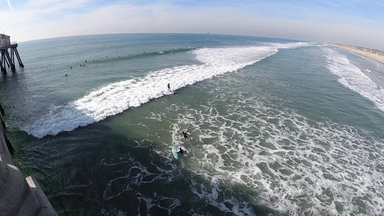 Wide surf view with pier supports, whitewater, and distant shoreline.