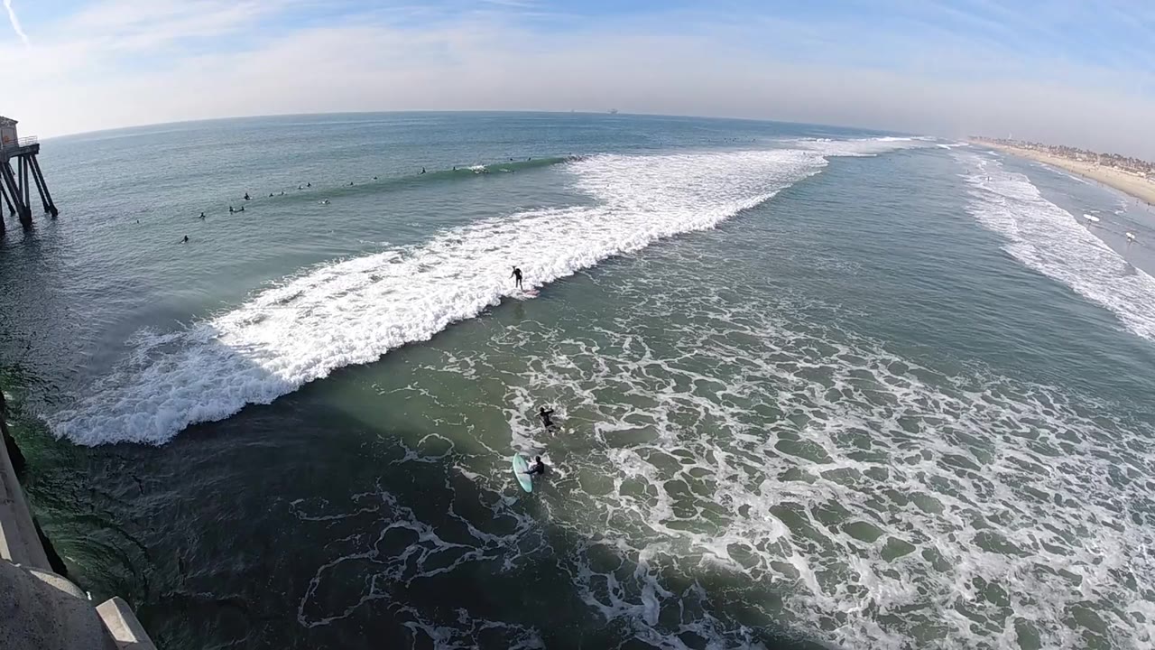 Huntington Beach surf spreads in a broad white band across blue-green water, with the beach and horizon beyond and small distant surfers or swimmers visible as dark specks.