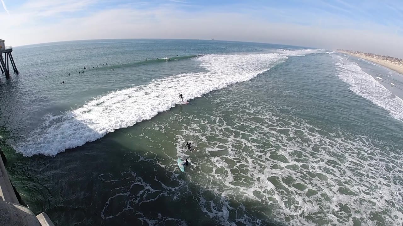 A wide Huntington Beach ocean view shows the wave crest stretched across the frame, the sandy shoreline receding to the right, and a dark pier support near the edge.