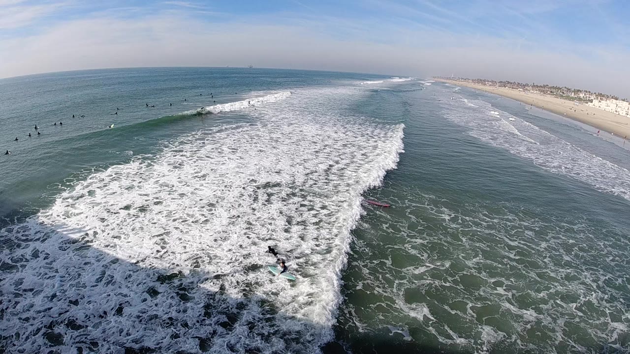 Elevated view over Huntington Beach surf at the start of the clip, with white foam filling the foreground and small surfers scattered beyond the breaking wave.