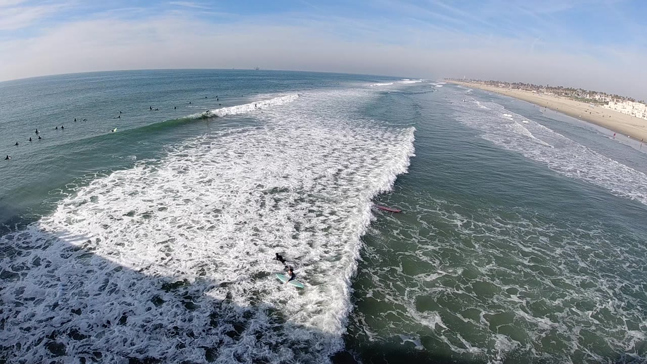 Huntington Beach aerial frame with the whitewater spreading lower in frame, surfers visible as dark figures offshore, and the sandy beach stretching into the distance on the right.