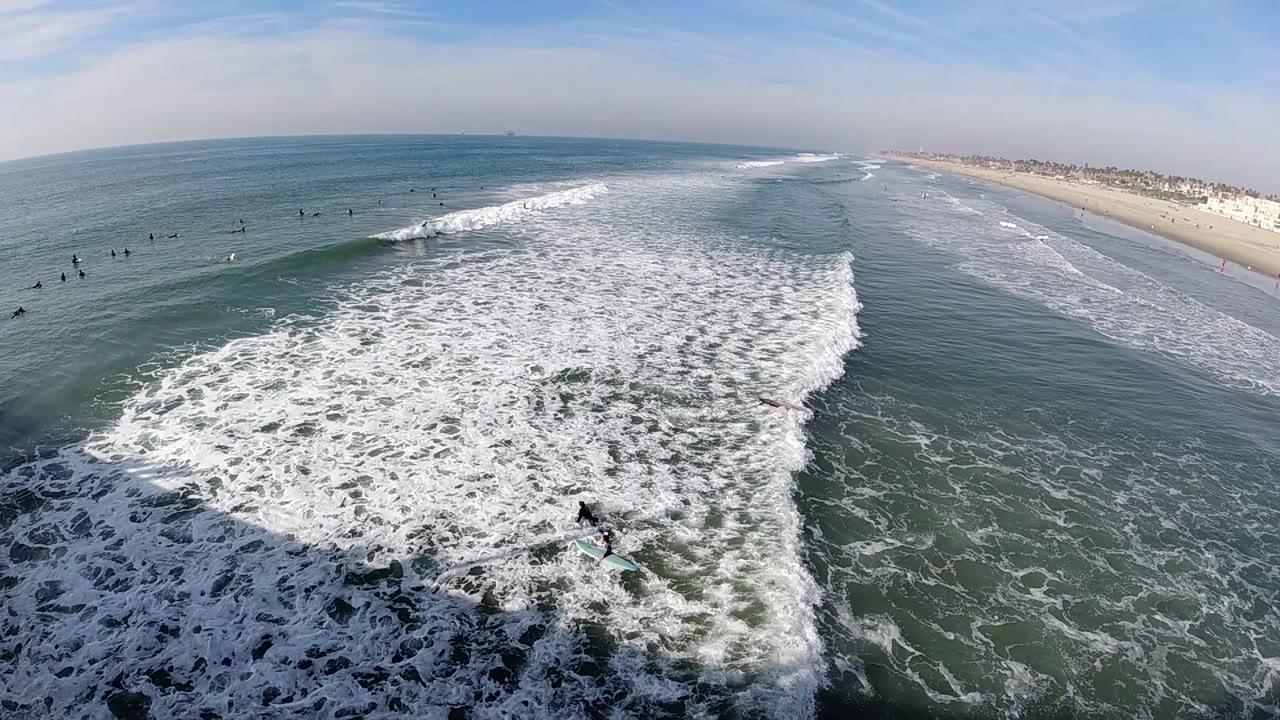 Aerial frame over Huntington Beach surf with white foam spreading across blue-green water, a sandy shoreline at right, and small distant surfers in the water.