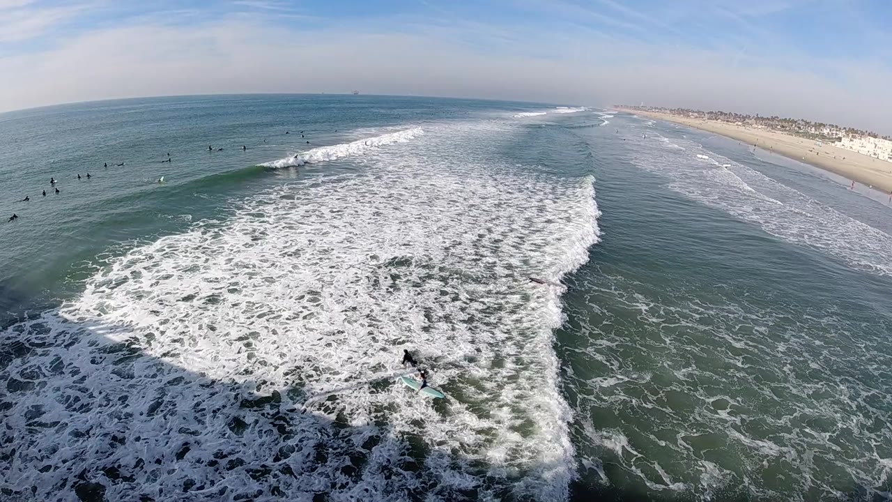 Aerial view of Huntington Beach with a wider sheet of whitewater crossing the foreground, distant surfers left of the break, and the beach running along the right side.