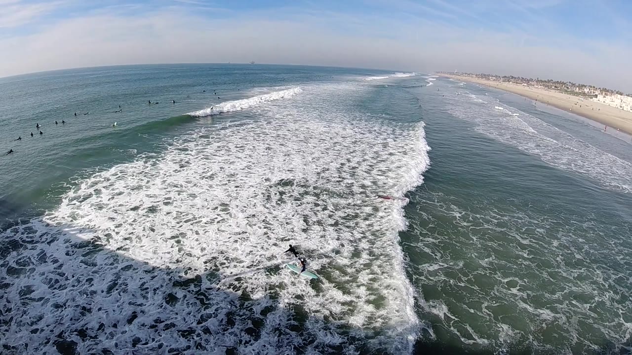 Aerial Huntington Beach surf scene with dense foam in the foreground, a green wave line behind it, tiny surfers scattered offshore, and the beach curving into the distance.