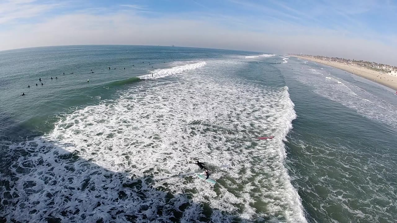 Aerial view over Huntington Beach with whitewater filling the foreground, surfers as small dark figures offshore, a sandy shoreline to the right, and no recognizable people.
