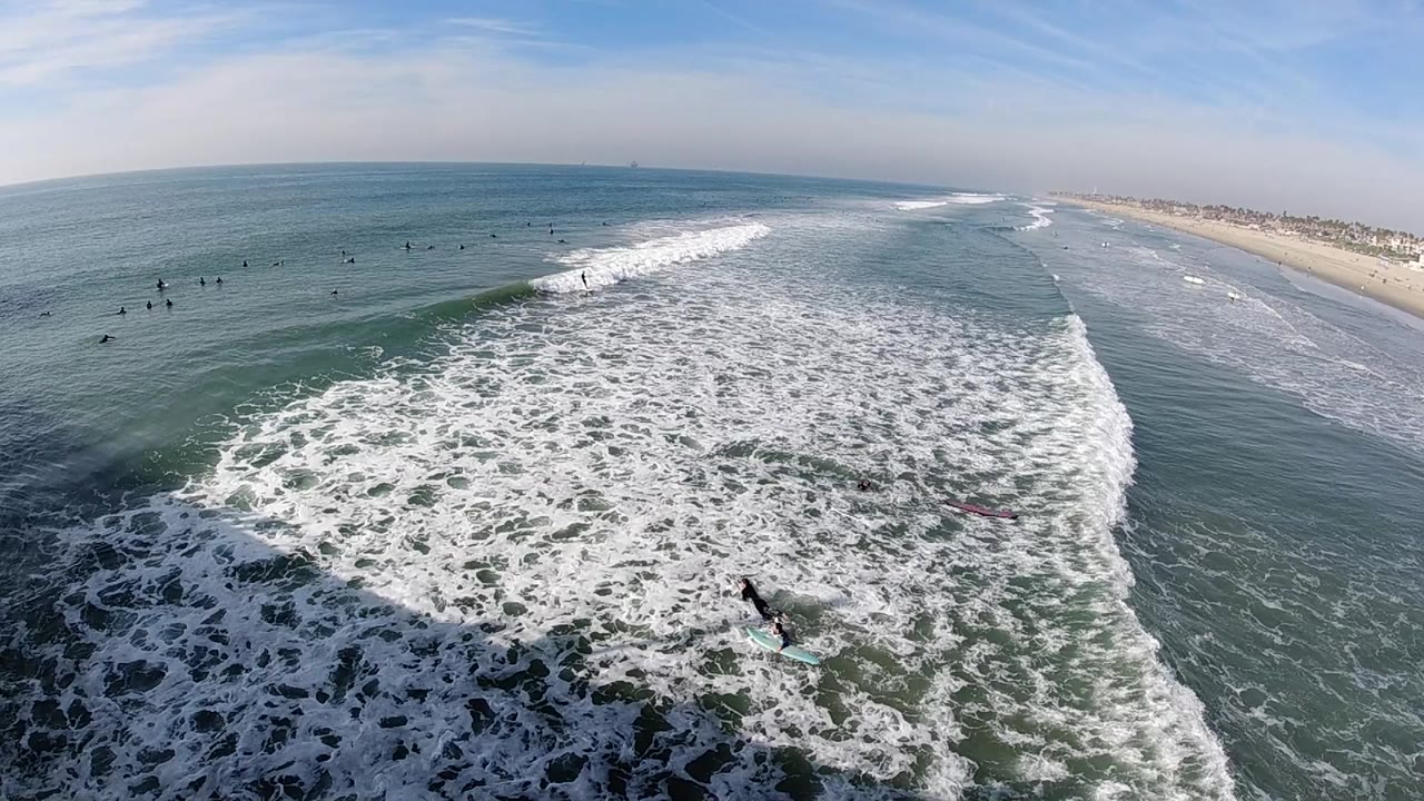 Elevated view over Huntington Beach surf at the start of the clip, with white foam filling the foreground, small surfers and swimmers offshore, and sandy shoreline to the right.