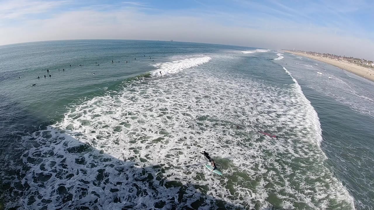Elevated Huntington Beach view at the start of the clip, with foamy surf spreading across green-blue water, small surfers offshore, sandy coastline in the distance, and no identifiable faces.