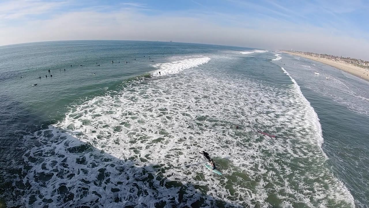 At 0:02, the elevated Huntington Beach view shows white water thick across the lower frame, calmer ocean beyond the break, tiny surfers waiting offshore, and the beach receding to the right.